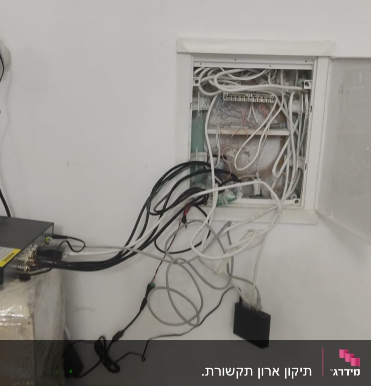 ארון תקשורת פתוח עם כבלים מחוברים ומכשיר רשת על שולחן.